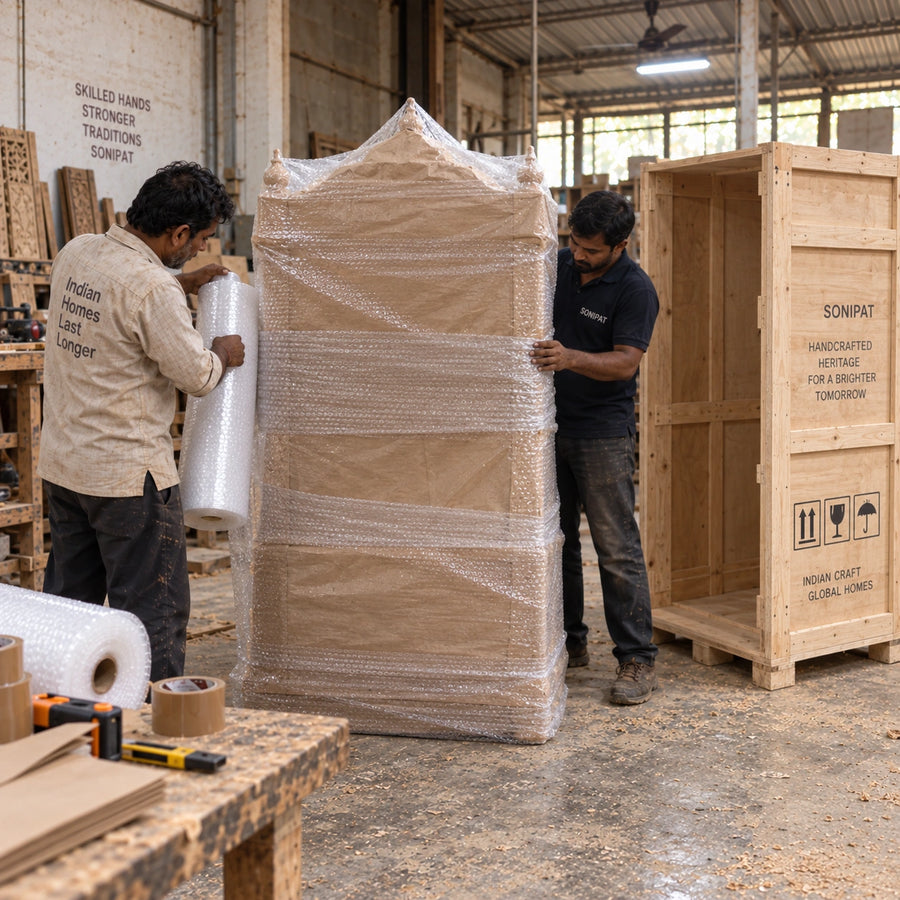 Two workers wrap a large wooden cabinet in bubble wrap inside a workshop, with a wooden crate and tools visible around them.