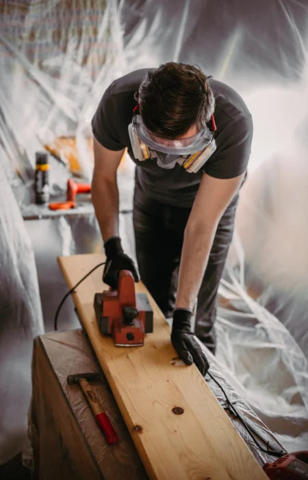 A carpenter wearing safety goggles and gloves using an electric sander to smooth a wooden plank in a workshop.