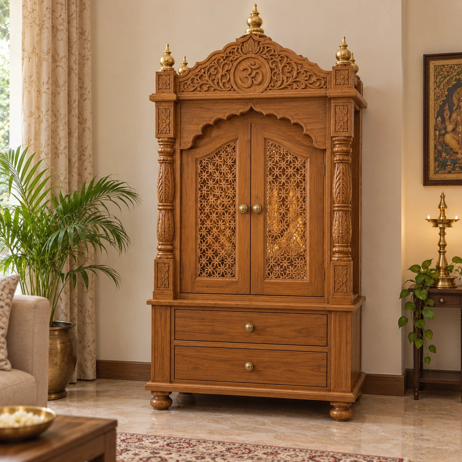 An ornate carved wooden pooja mandir from DZYN Furnitures with intricate floral detailing, two doors, and two drawers, shown in a styled living room setting.